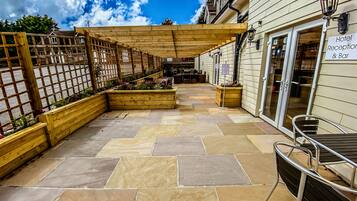 Un patio con suelo de piedra, jardineras de madera con flores, una pérgola y una zona de bar con un letrero que indica recepción de hotel y bar.