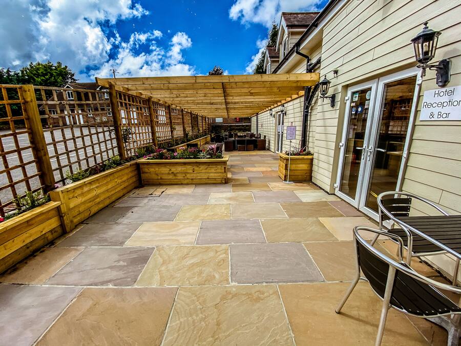 Uma área de pátio com piso de pedra, vasos de madeira com flores, uma pérgola e um espaço de bar com uma placa indicando a recepção do hotel e bar.