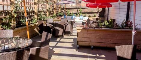 Une terrasse extérieure avec des meubles en osier, des parasols rouges et un jardinier en briques.