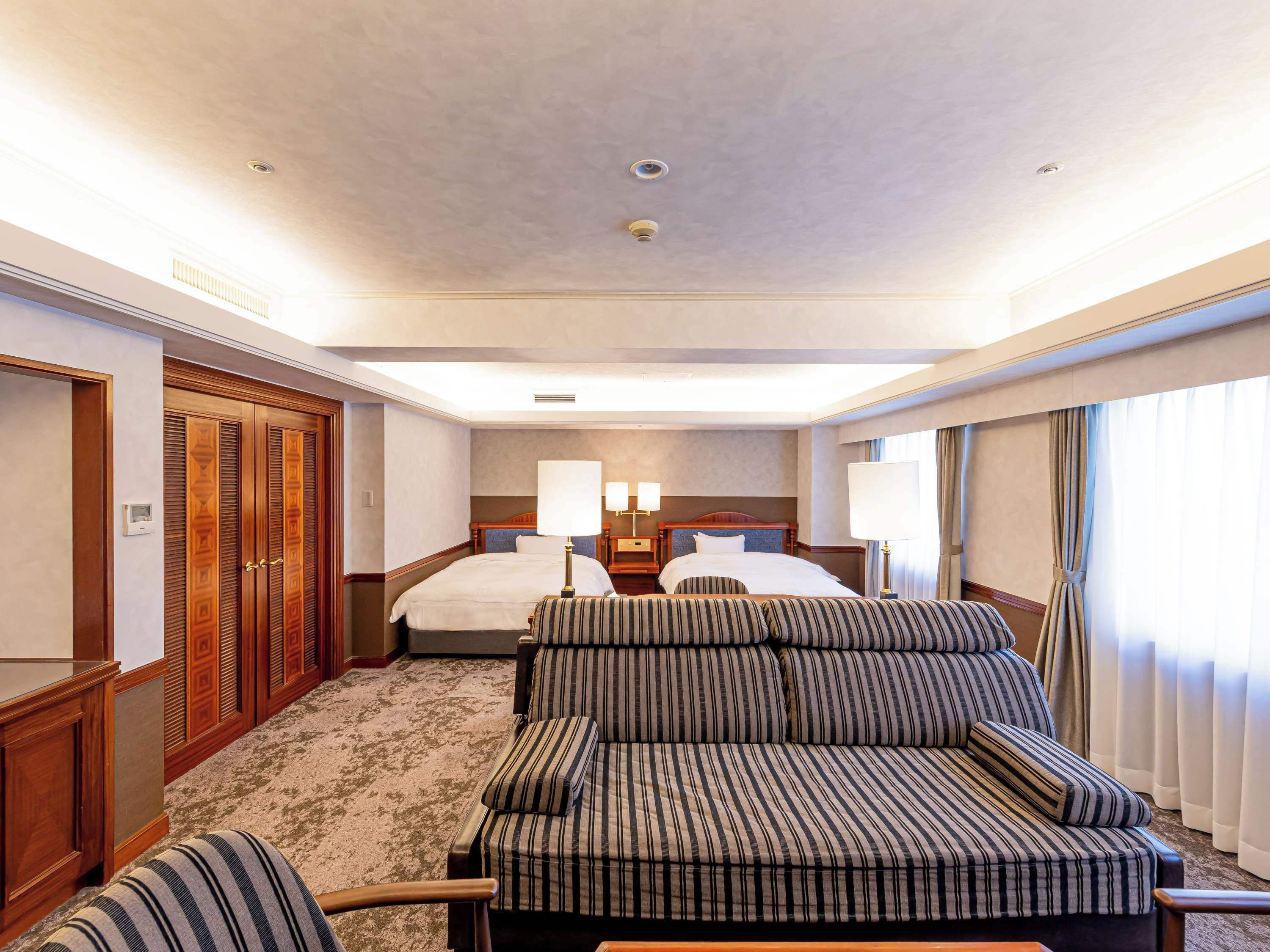 A hotel room with two beds, a sofa, a desk, and a large window with curtains.