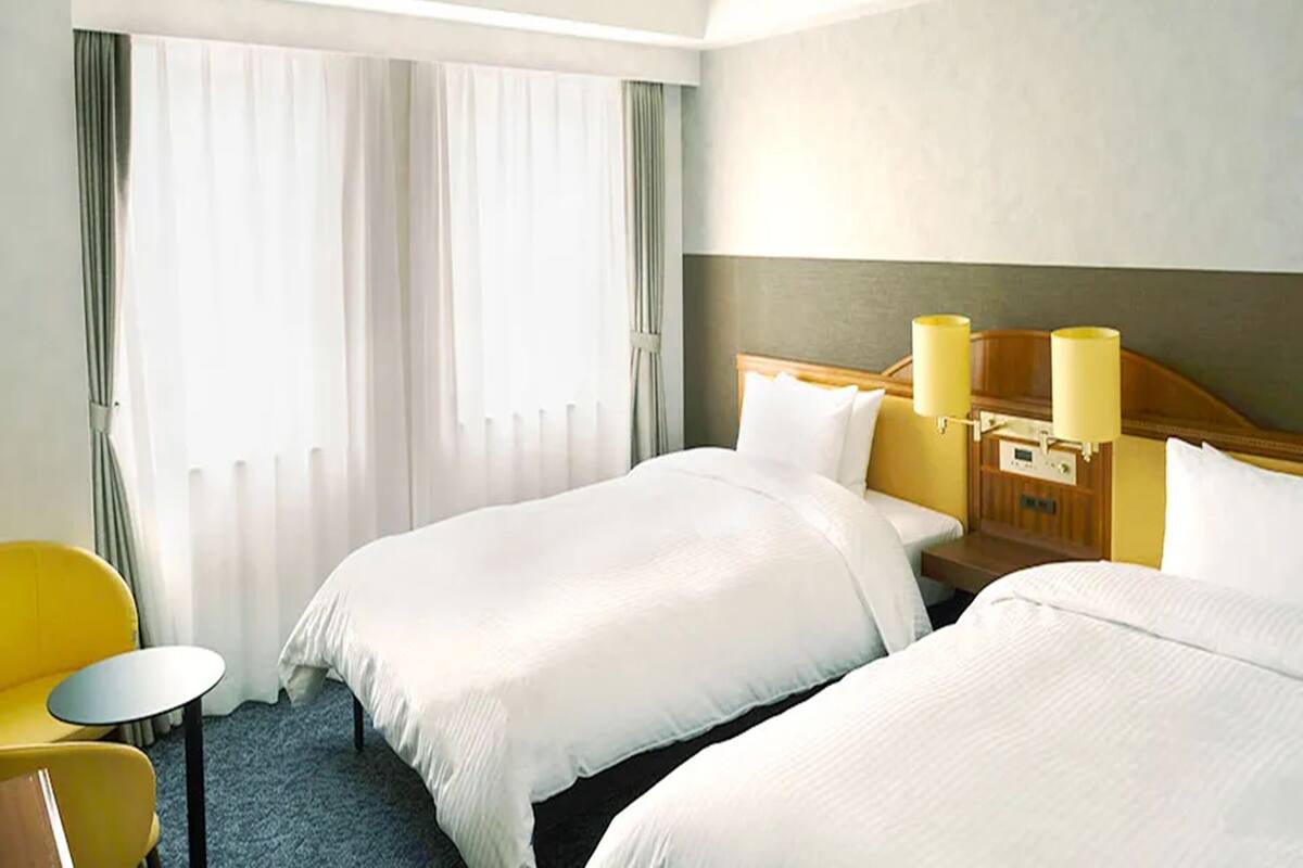A hotel room with two beds, a small table, a chair, and a window with curtains.