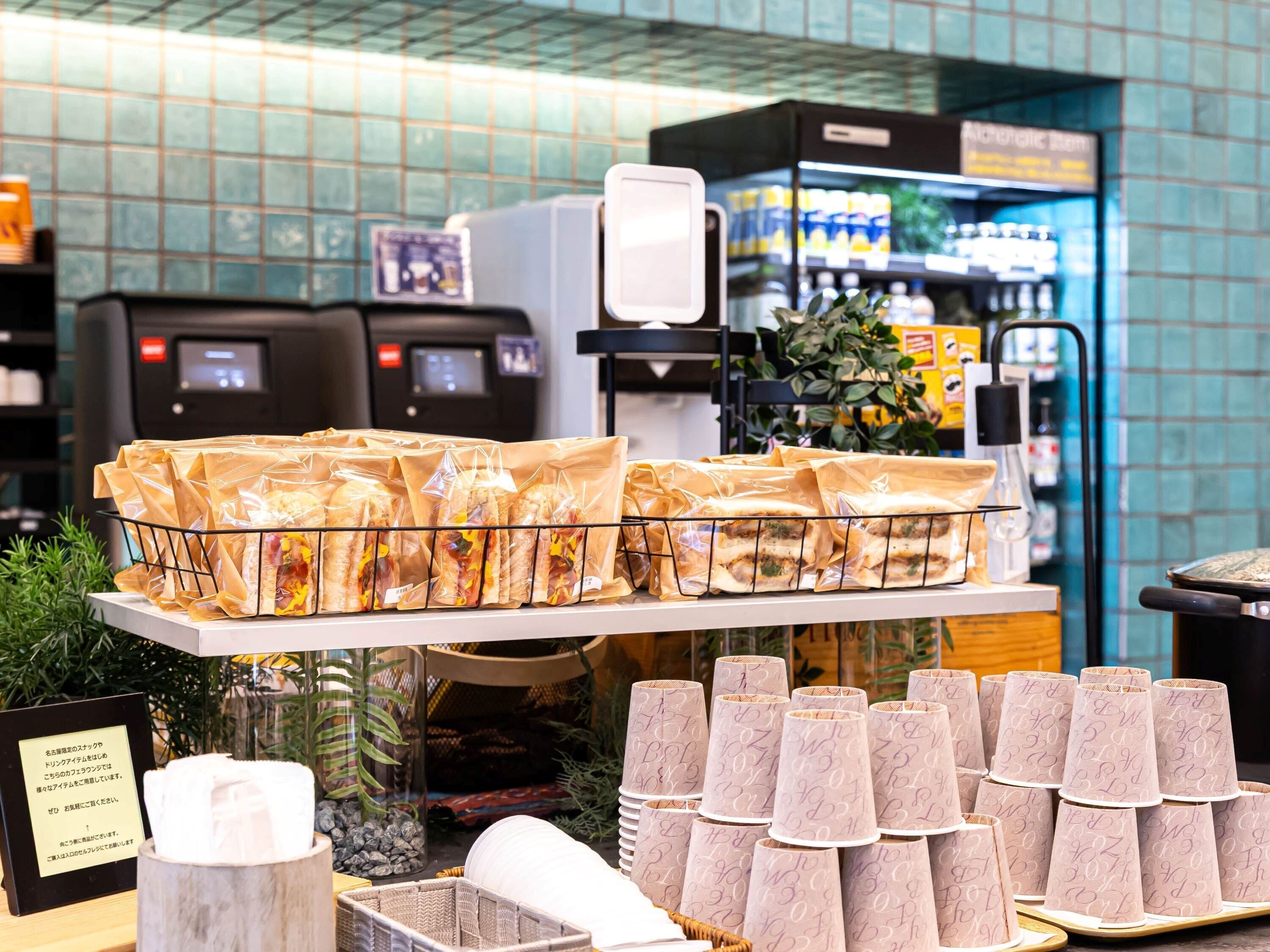 A display of packaged food items and paper cups with a sign, in a store setting with a tiled wall and a coffee machine in the background.