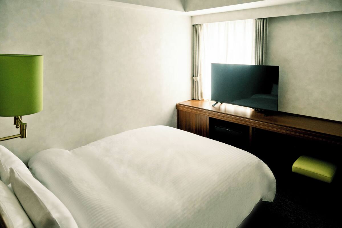 A hotel room with a bed, a television, a desk, and a green lamp.