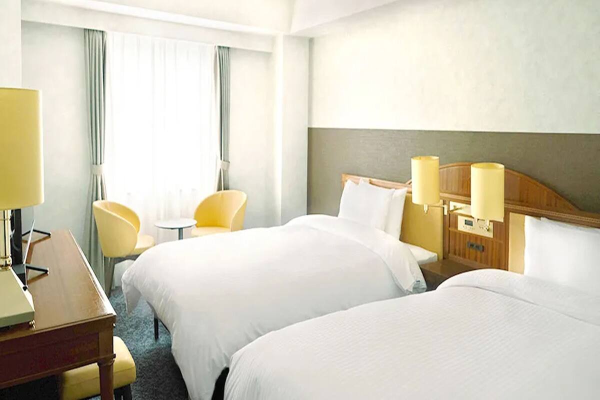 A hotel room with two beds, a desk, a chair, and a lamp.