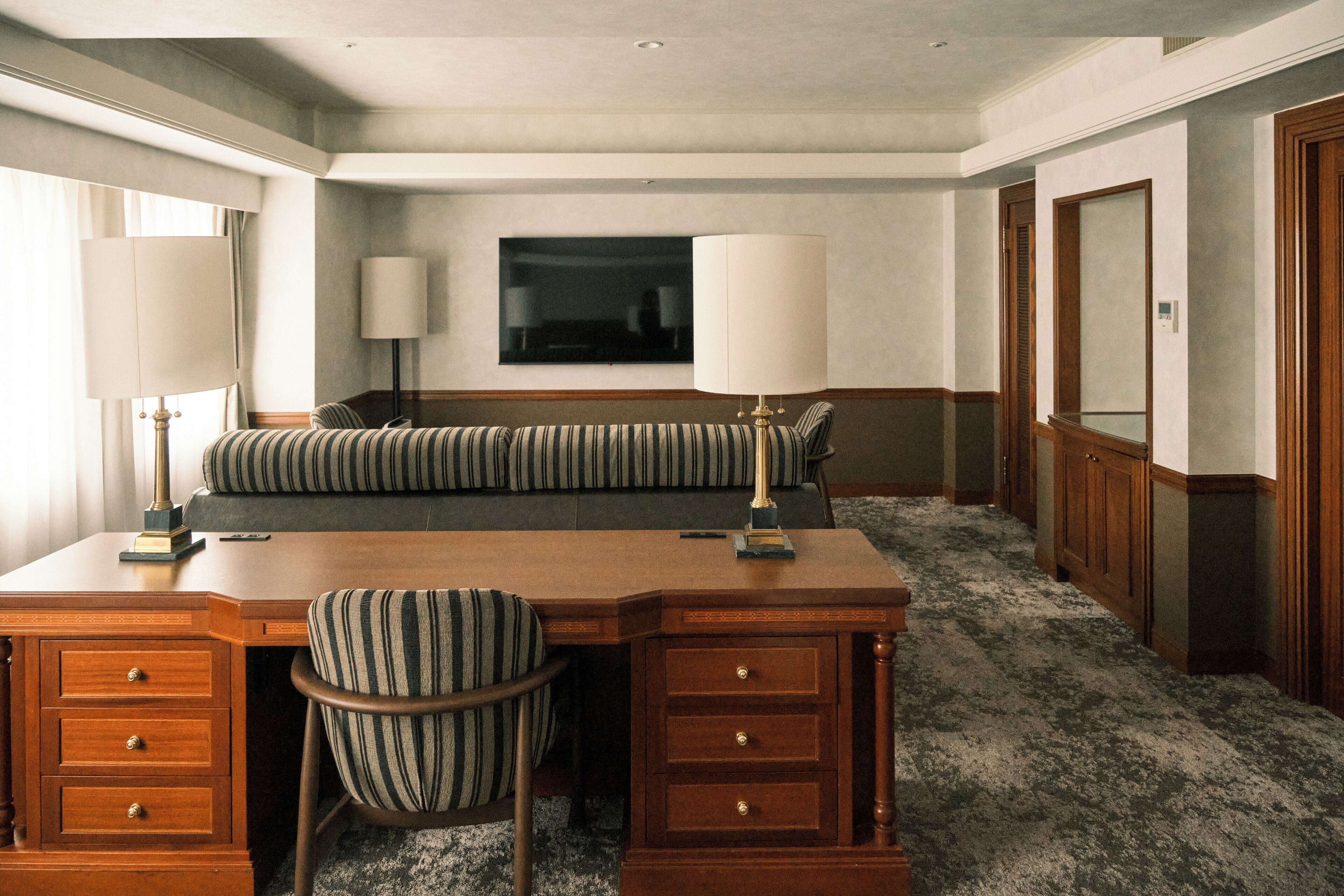 A hotel room with a wooden desk, two lamps, a striped sofa, a television, and a wooden wardrobe.