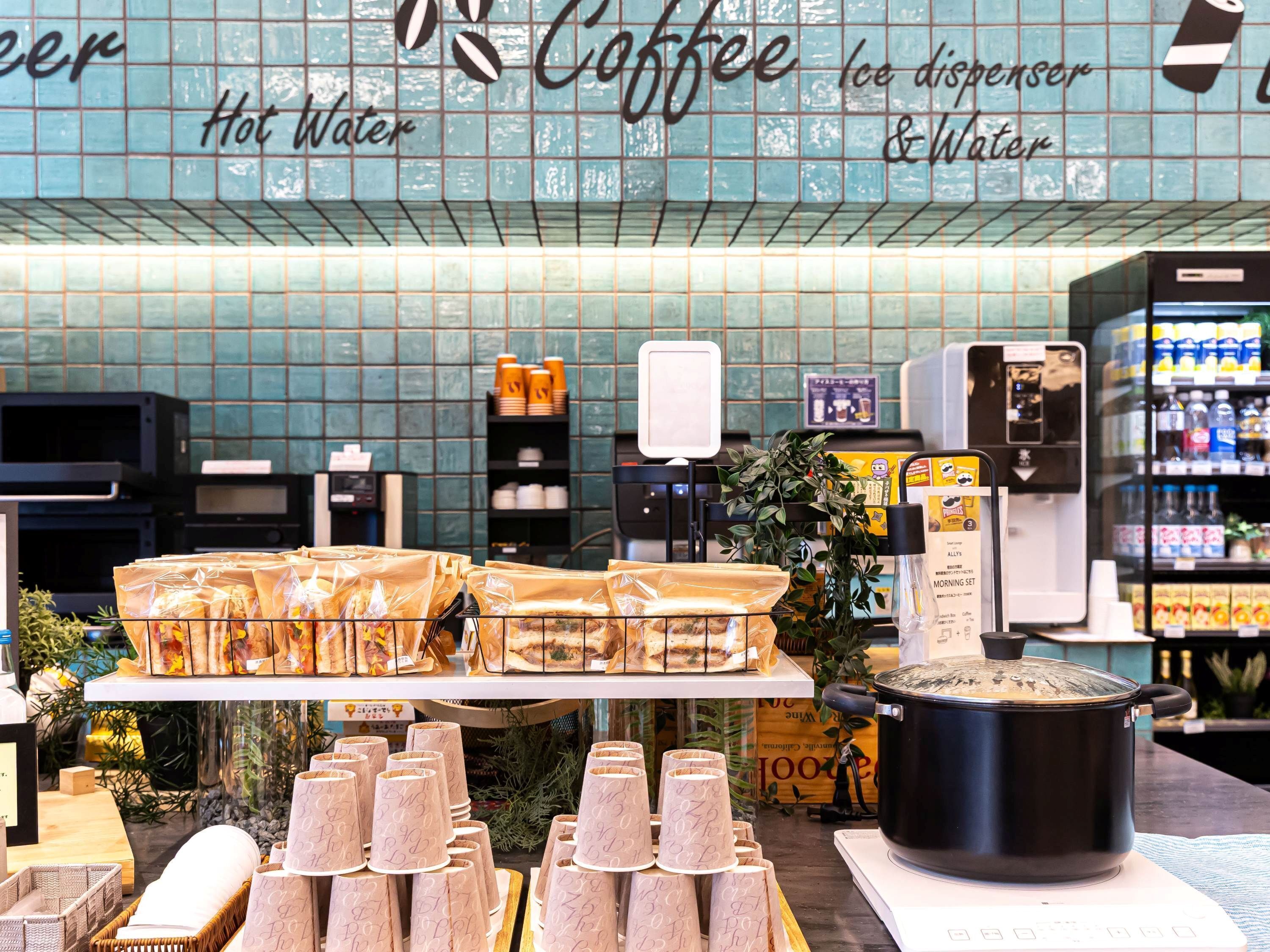 Interno di una caffetteria con una macchina da caffè, un dispenser di acqua calda e una selezione di tazze e sacchetti di caffè.