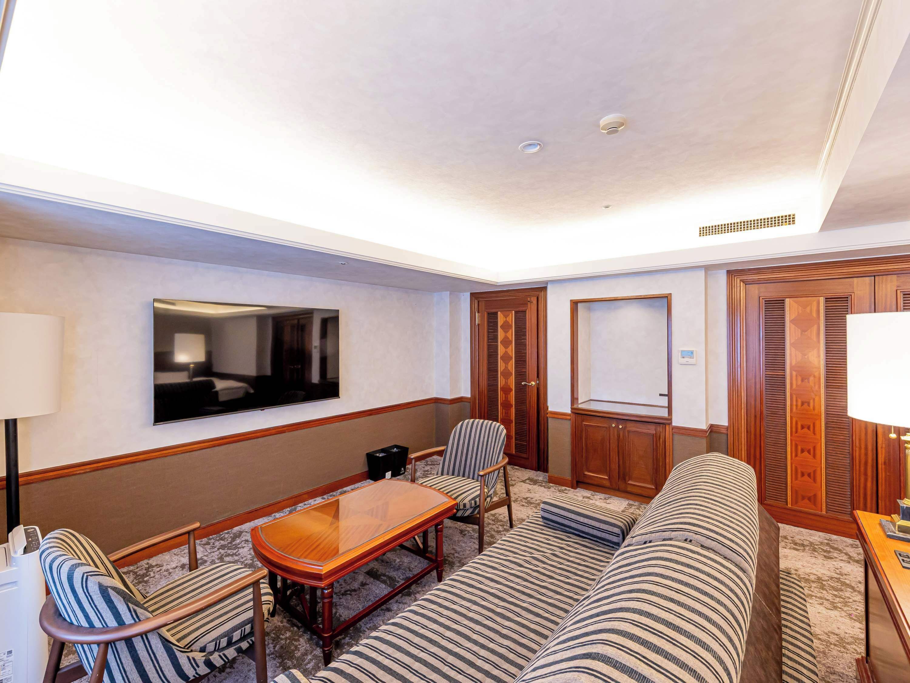 A hotel room with a sofa, a wooden coffee table, a TV mounted on the wall, a wooden wardrobe, and a lamp.