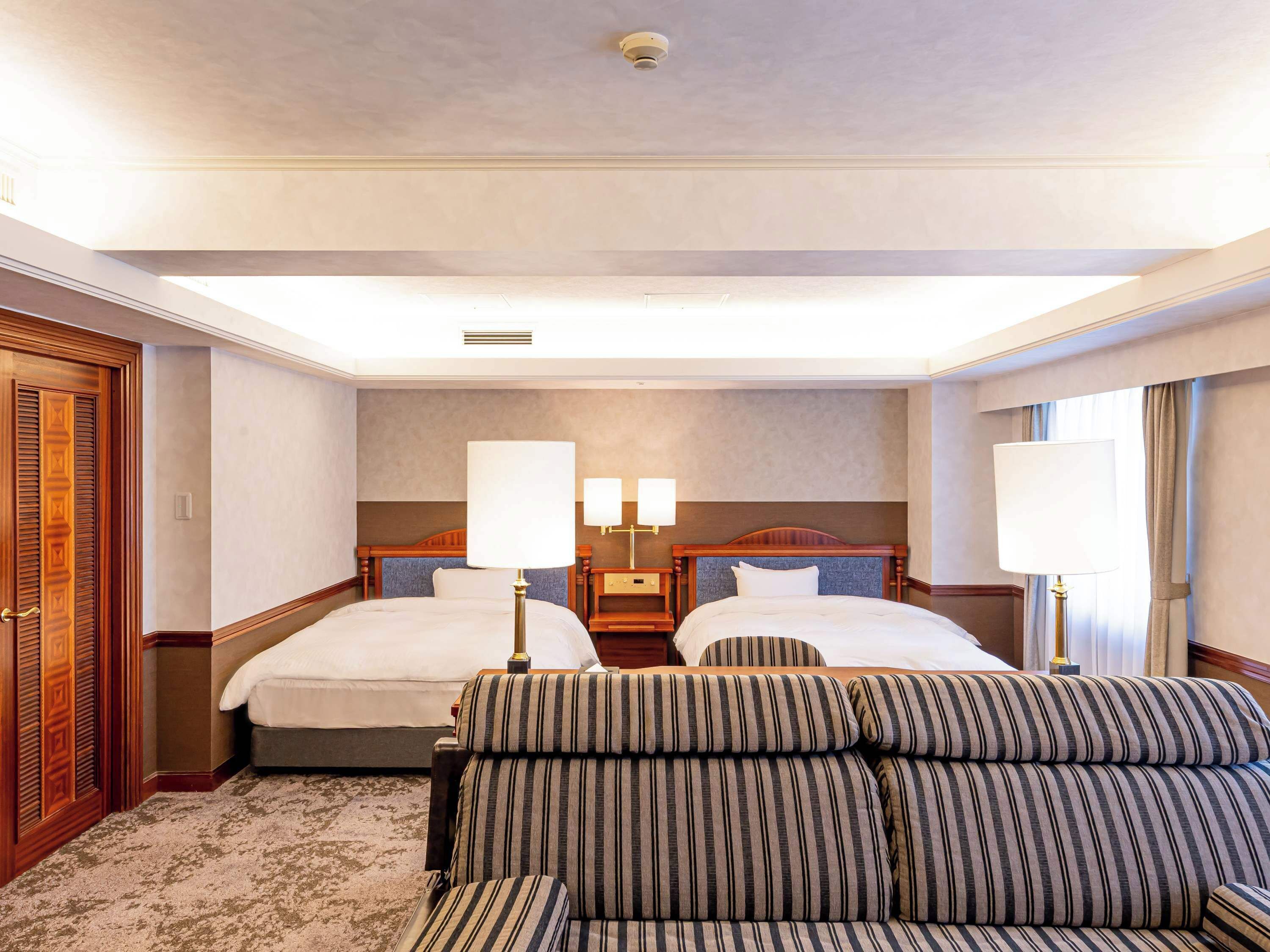 Habitación de hotel con dos camas, un sofá, un armario de madera y un detector de humo en el techo.