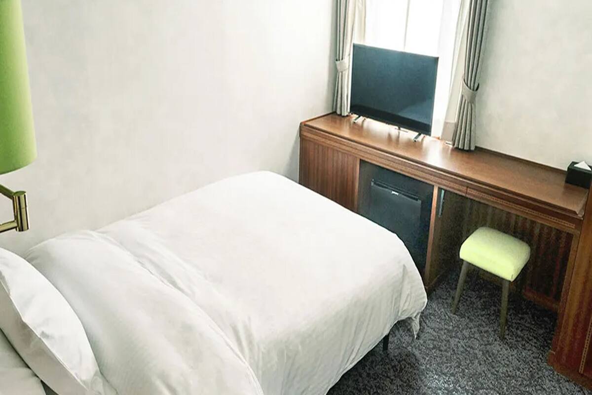A hotel room with a bed, a desk with a television, a chair, and a lamp.