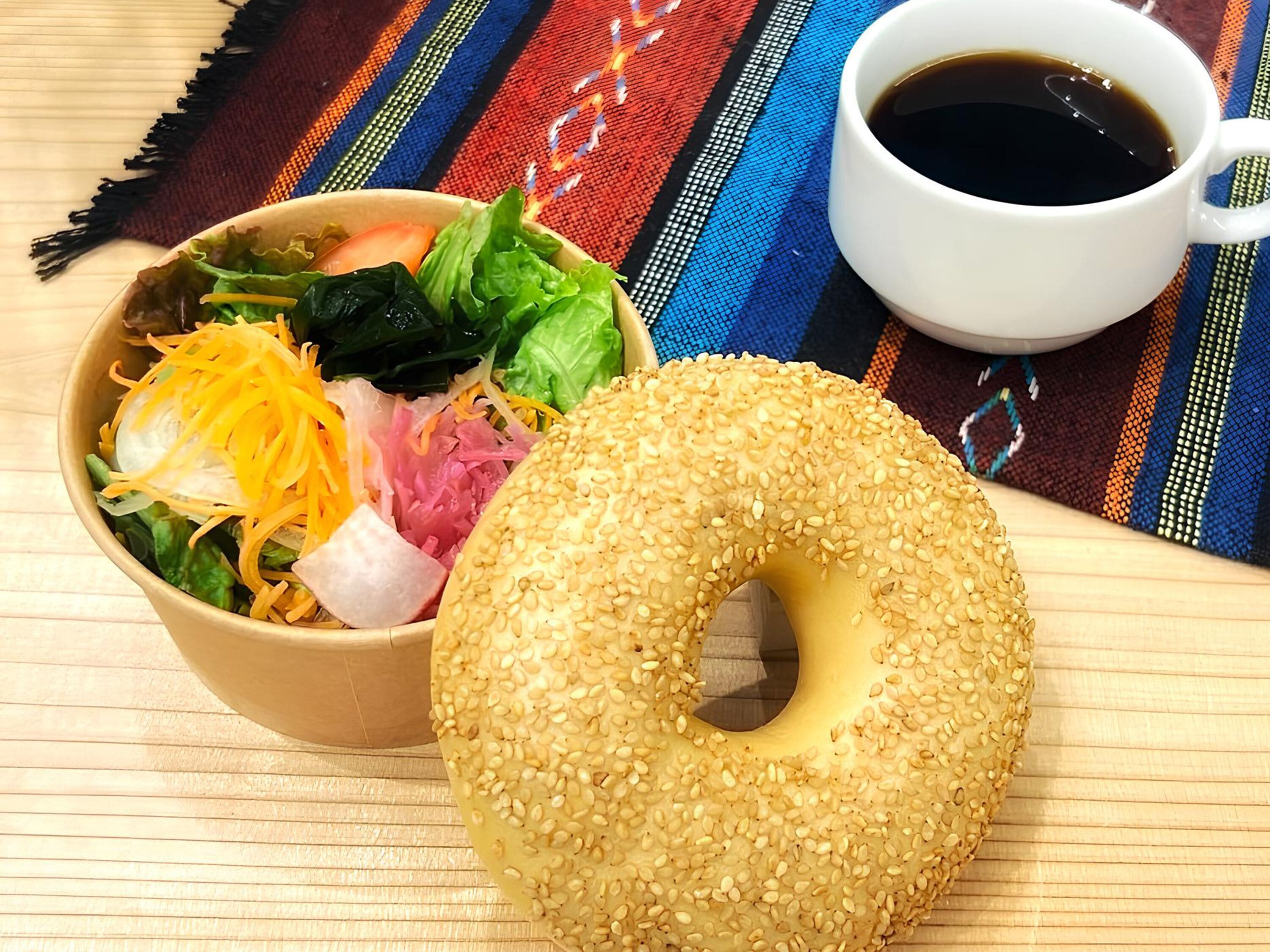 A meal with a sesame seed bagel, a salad, and a cup of coffee on a table with a colorful woven placemat.