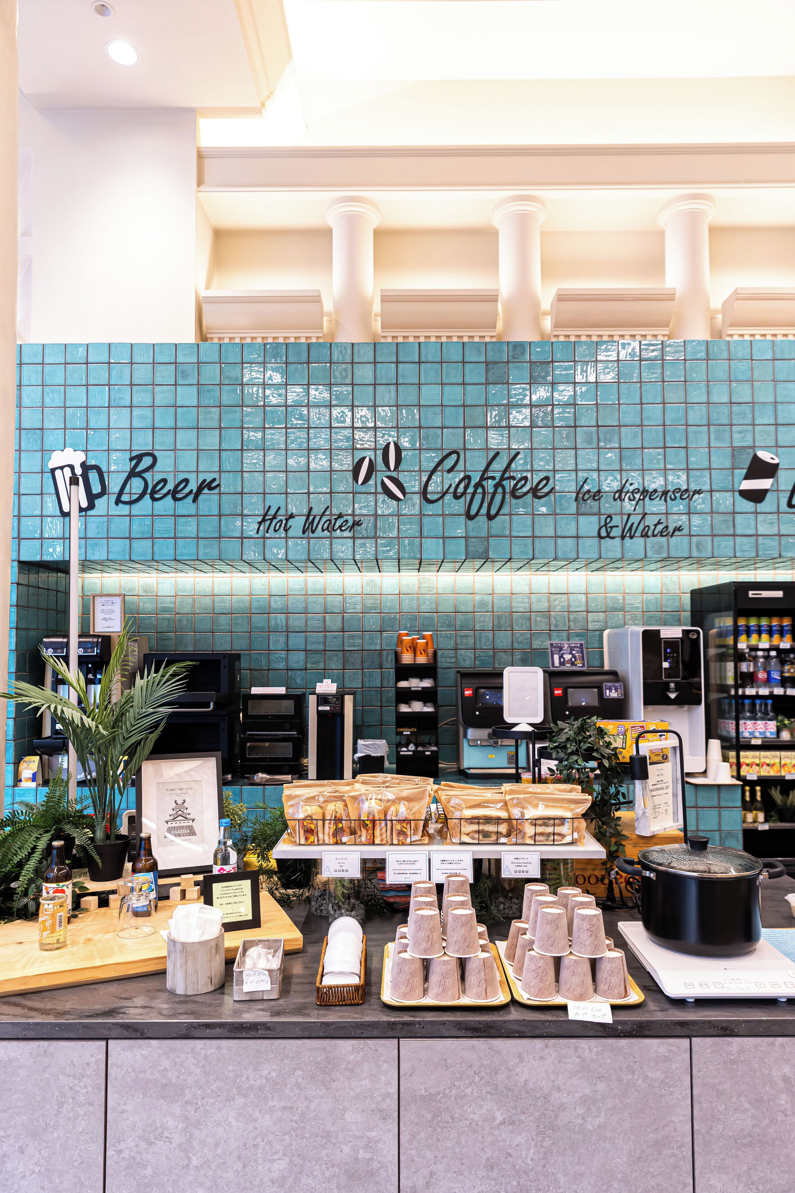 Café com parede de azulejos azuis e um letreiro que diz 'Cerveja Água Quente Café'. Há máquinas de café, uma variedade de copos de papel e uma panela grande em um fogão portátil.