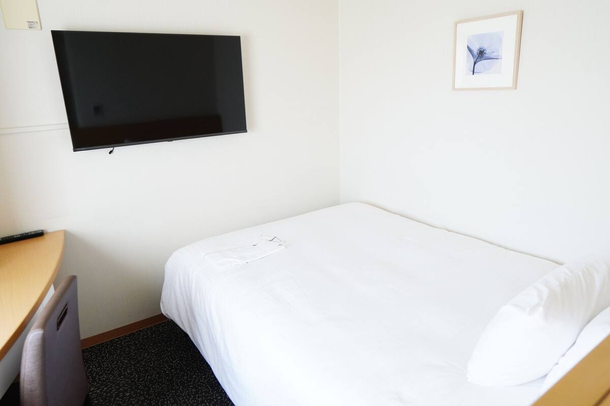 A hotel room with a bed, a desk, a chair, a television mounted on the wall, and a framed picture on the wall.