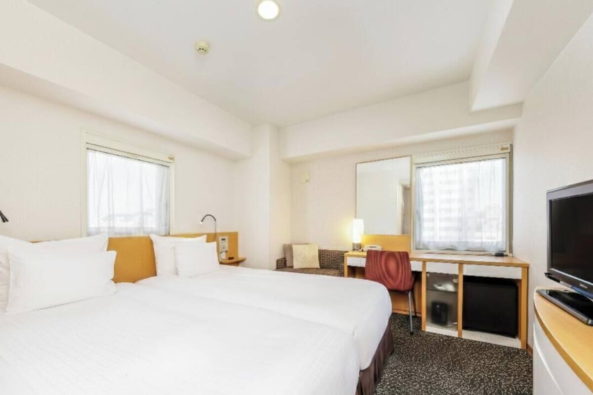 A hotel room with a large bed, a desk with a chair, a television, and two windows with curtains.