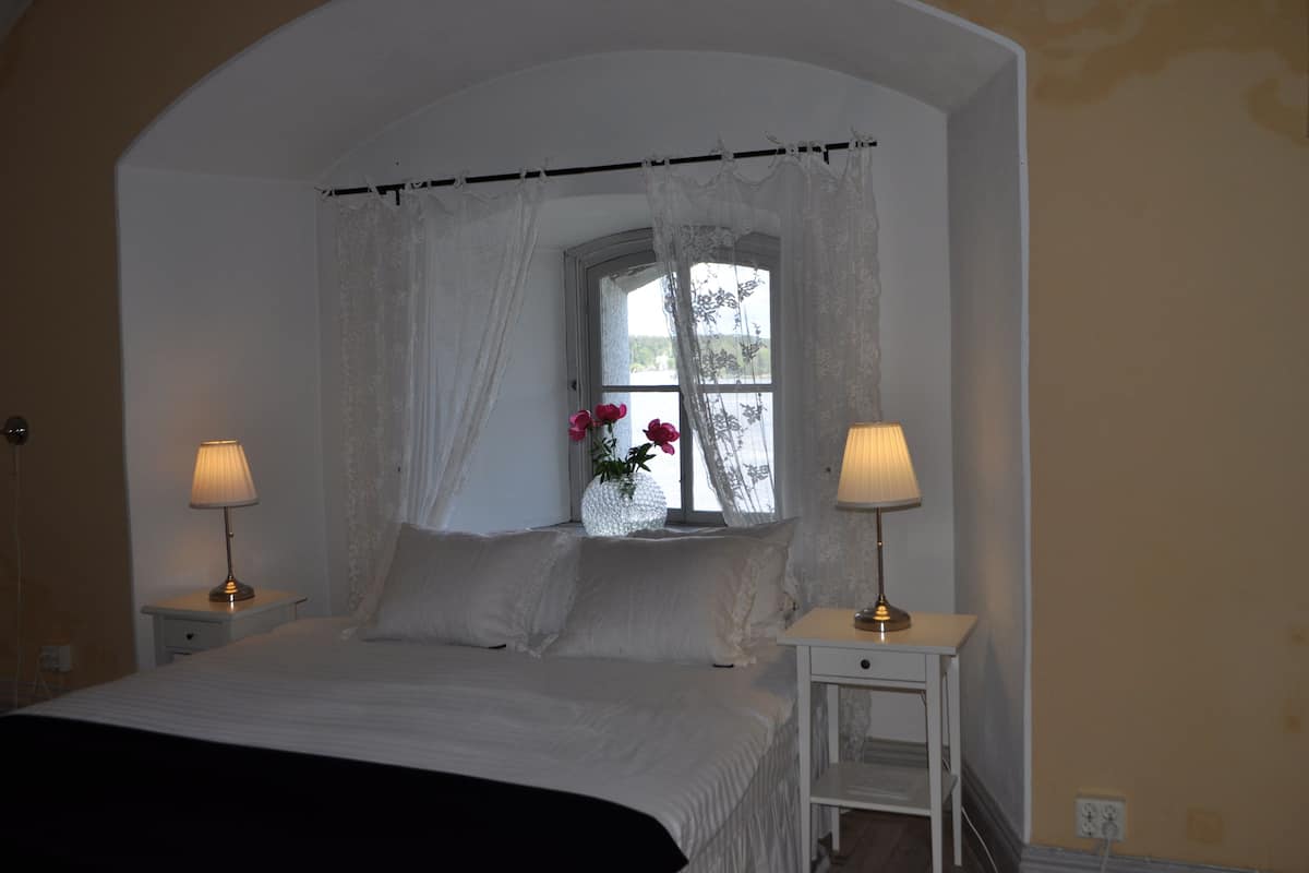 Ein Schlafzimmer mit einem Bett, zwei Nachttischen mit Lampen, einem Fenster mit Spitzenvorhängen und einer Vase mit Blumen.