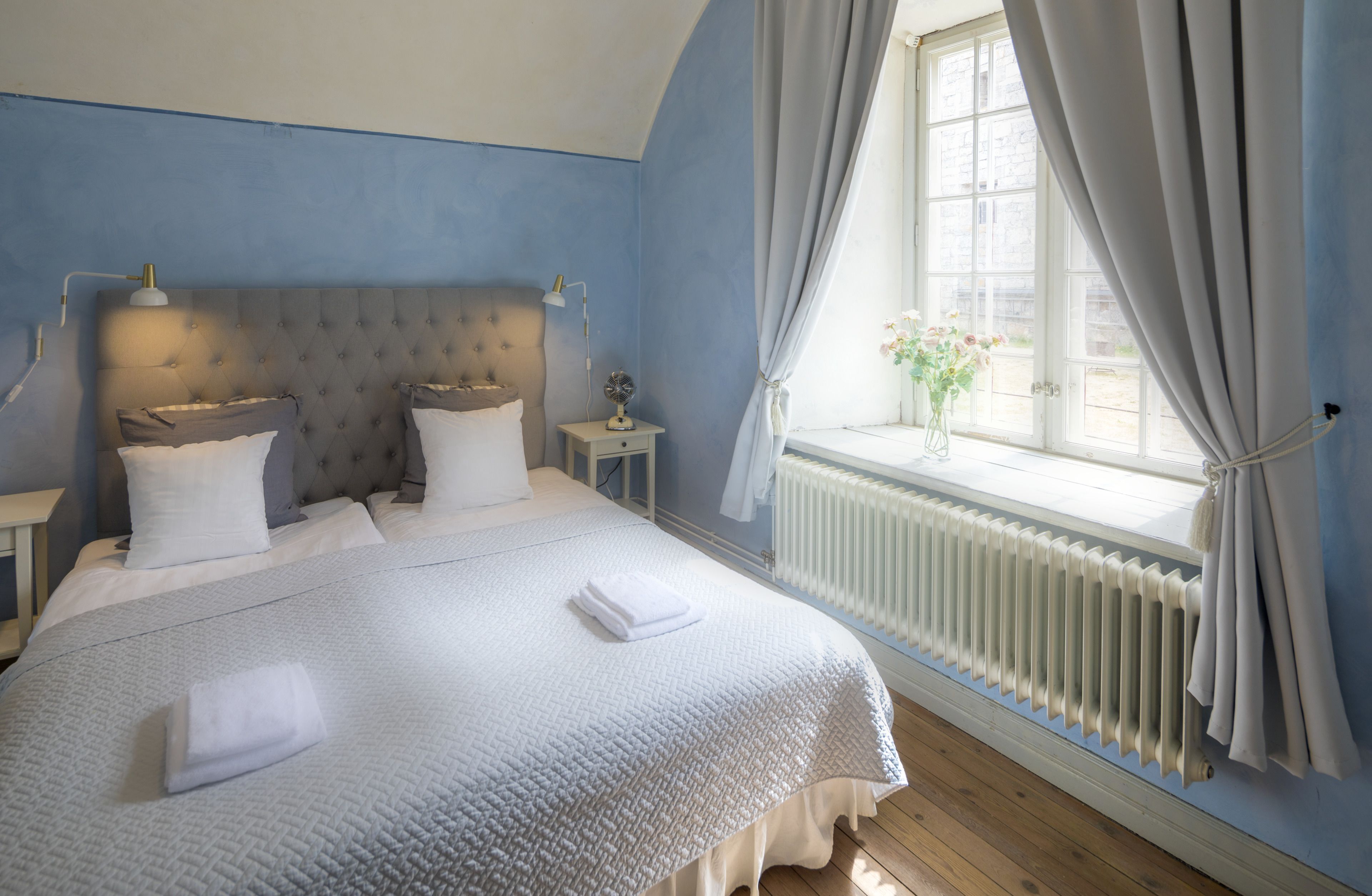 A neatly made bed with white linens and a folded towel, a nightstand with a lamp, and a window with curtains.