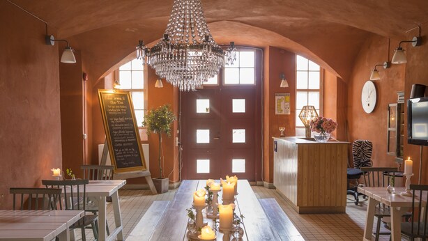 Un interior de restaurante con mesas de madera, una araña y una pizarra con el menú.