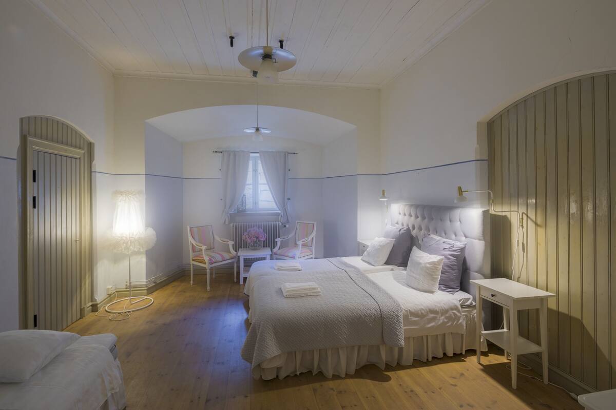 Ein Schlafzimmer mit einem großen Bett, einem Sessel, einem Nachttisch und einem Fenster mit Vorhängen.
