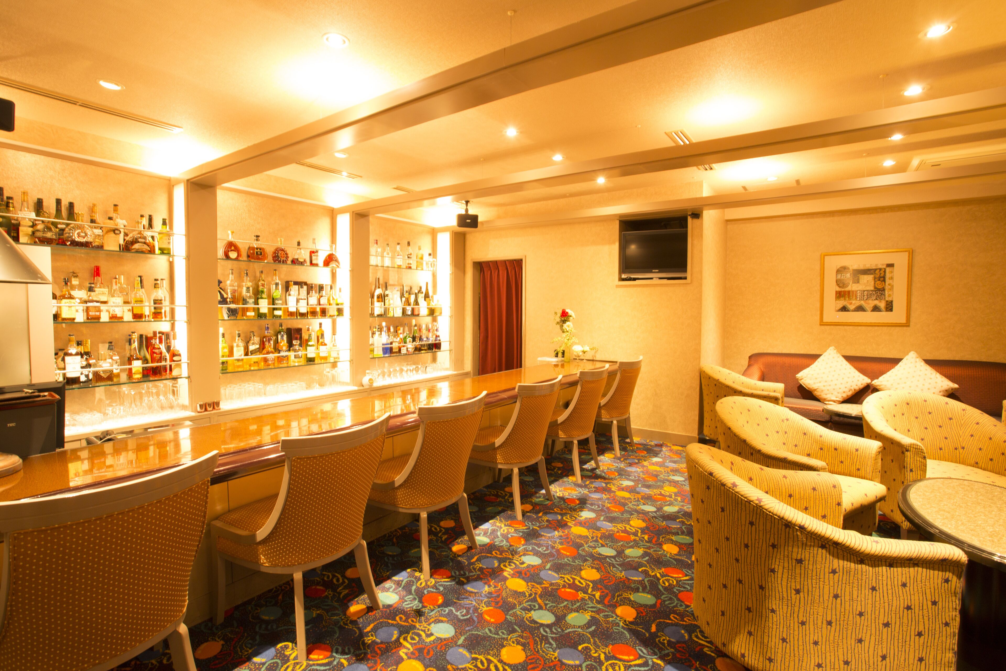 A bar area with multiple seating options, a variety of bottles on shelves, and a television mounted on the wall.