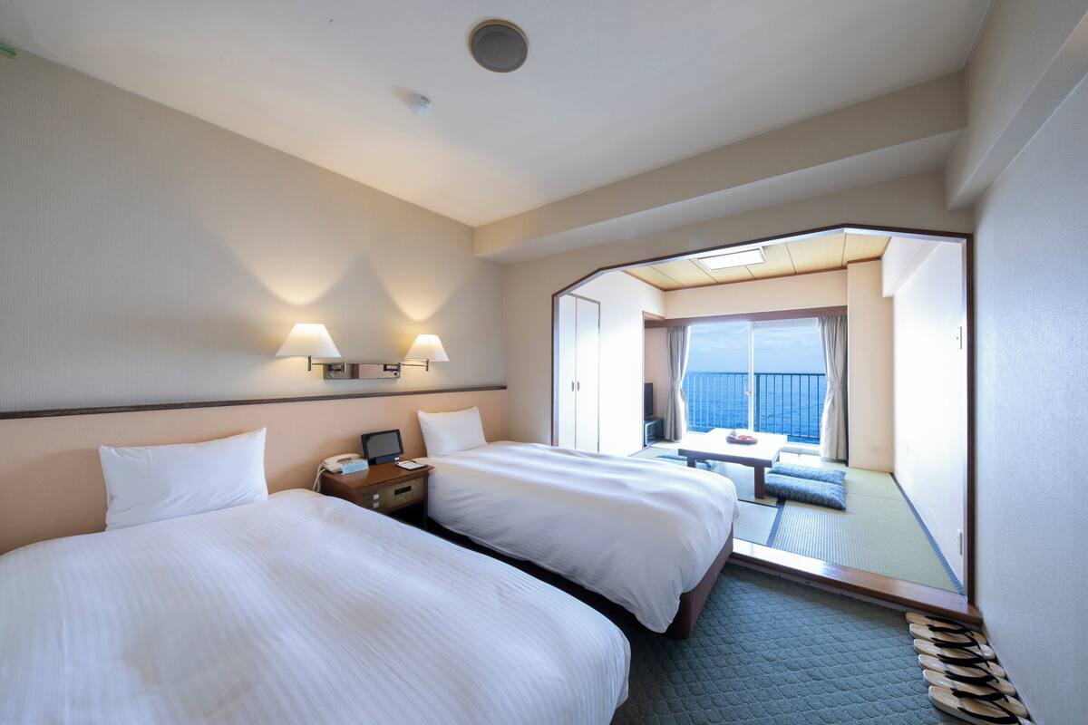Habitación de hotel con dos camas, un escritorio con lámpara y vista al mar a través de un ventanal grande.