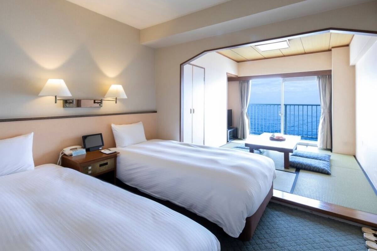 Habitación de hotel con dos camas, un escritorio, una silla y un balcón con vista al mar.