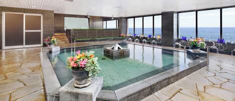 A large indoor swimming pool with a view of the sea, stone flooring, and seating areas with drinks.