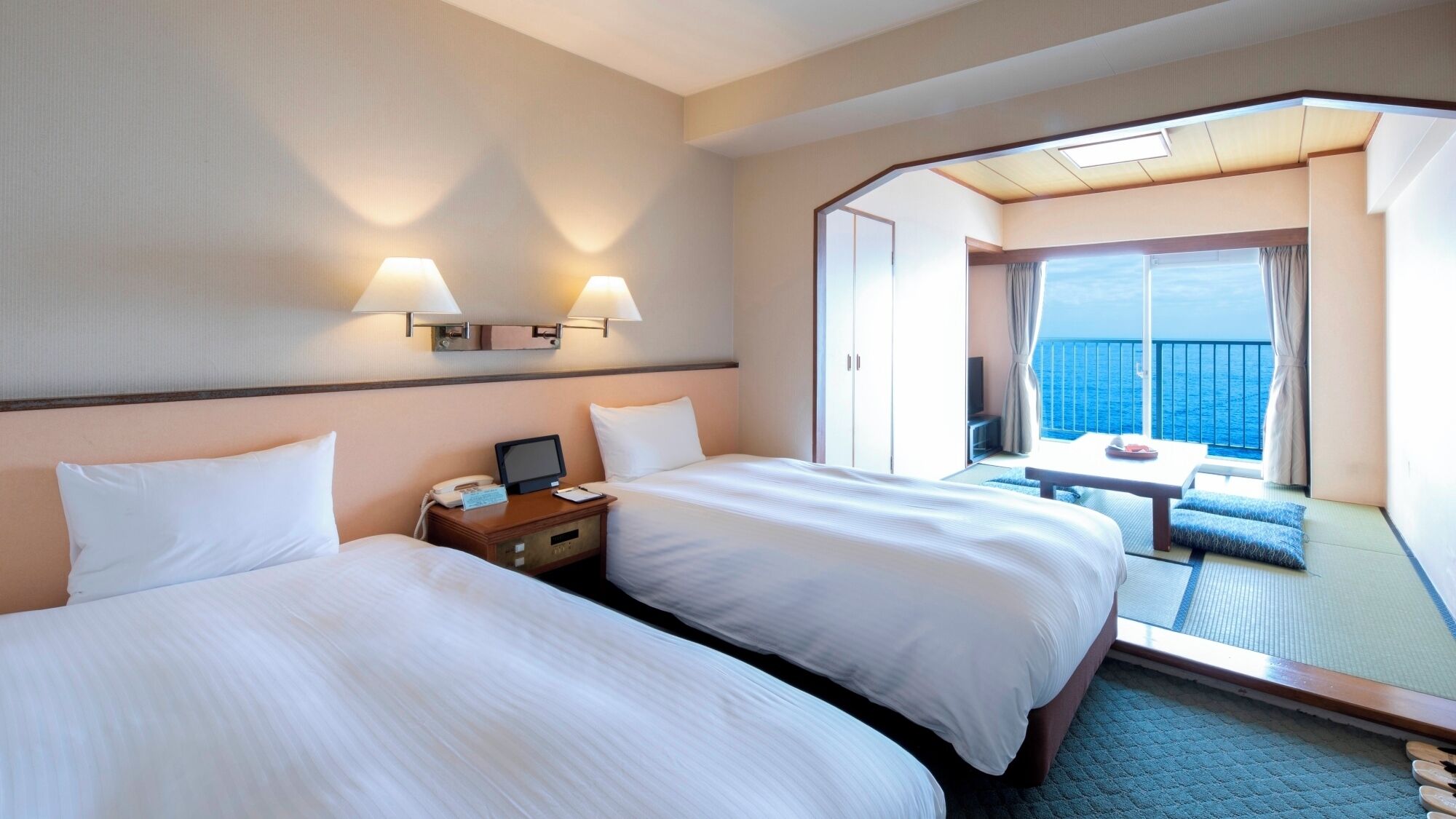 Habitació d'hotel amb dos llits, una escriptori amb un telèfon i un ordinador, una taula petita amb una cadira i un balcó amb vista al mar.