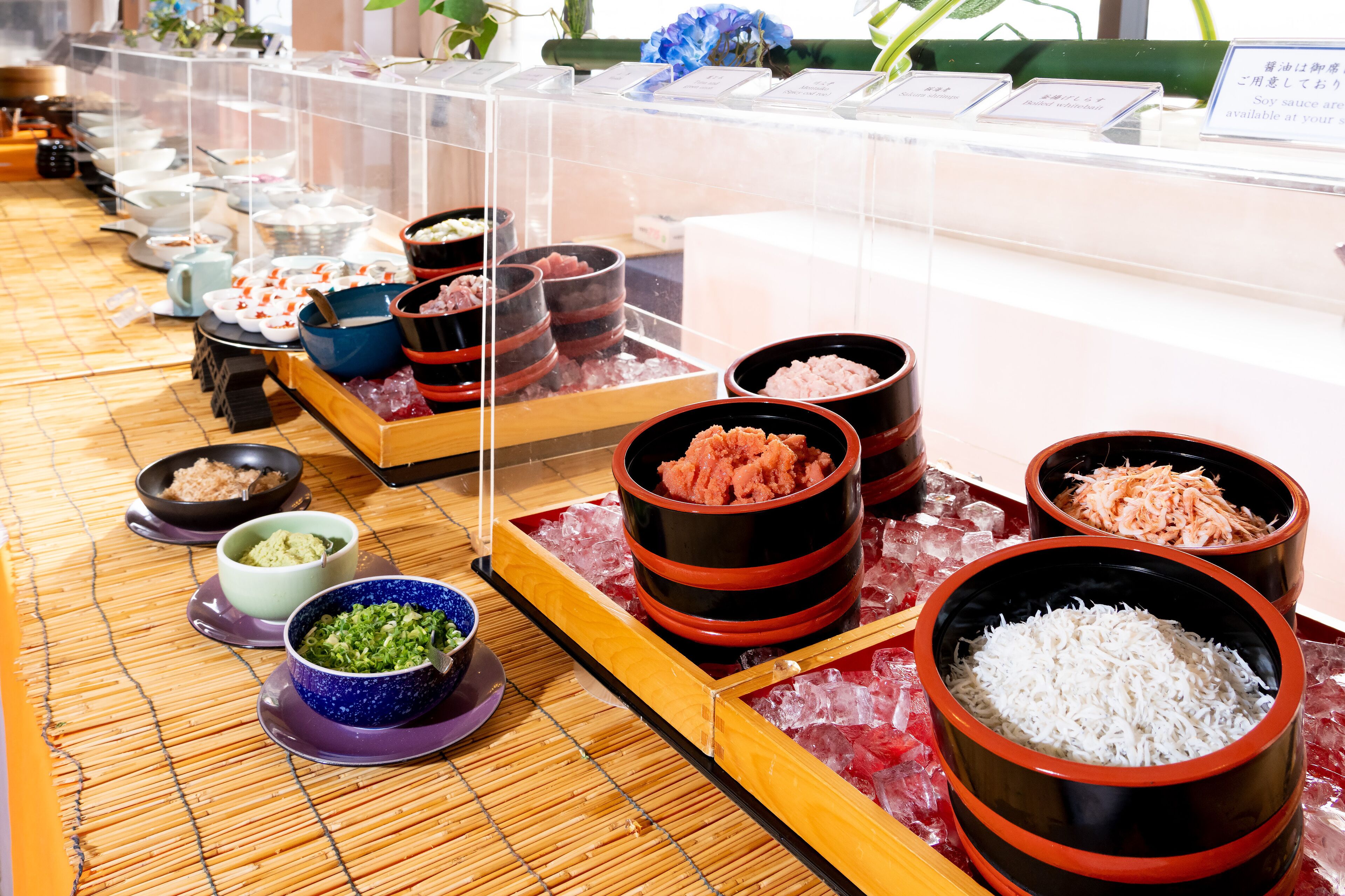 A buffet with various dishes displayed in black and red bowls on ice, with a bamboo mat on the floor.