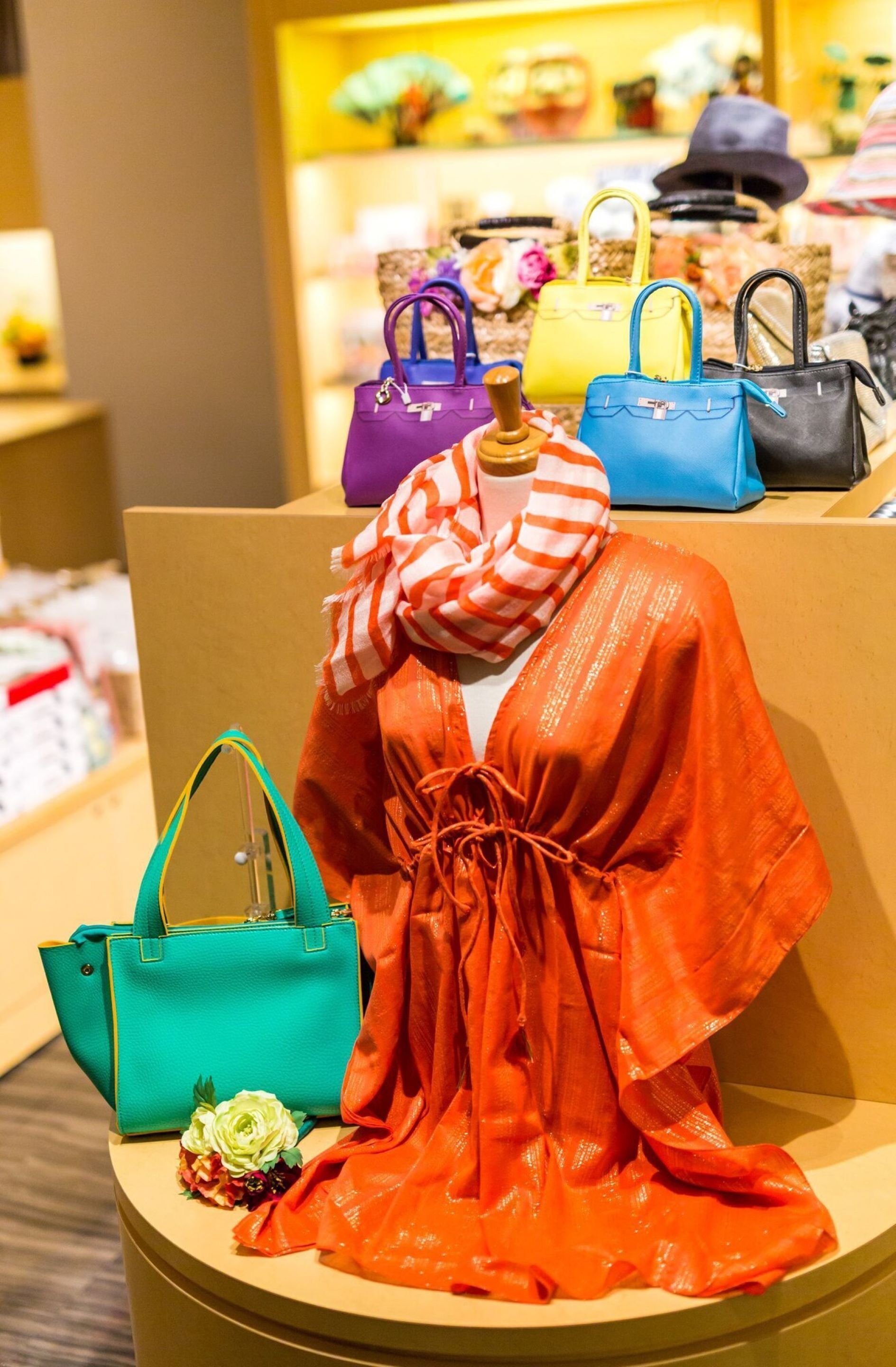 A mannequin dressed in an orange dress with a bow, surrounded by various handbags on a display stand.