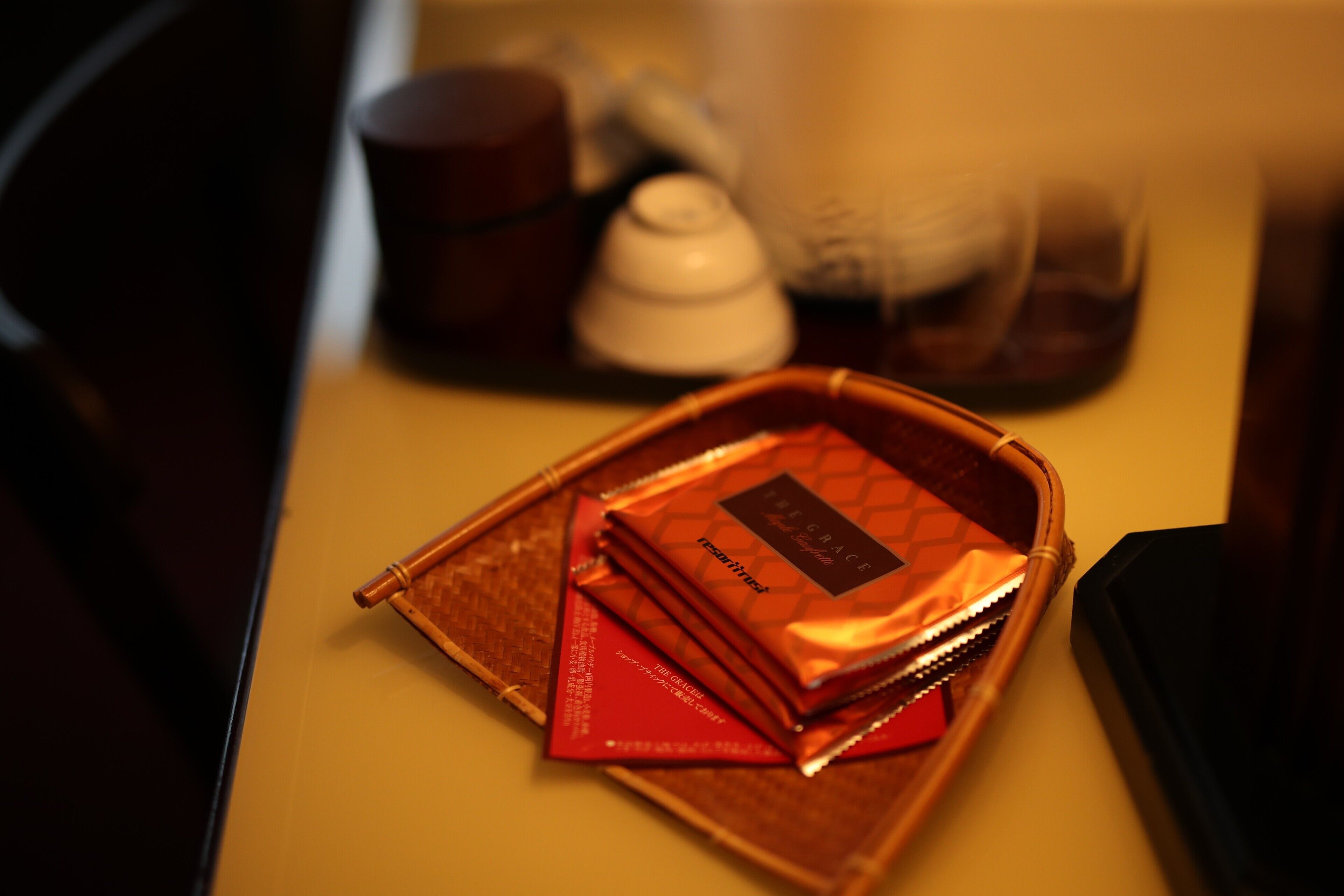 Una bandeja con una cesta trenzada marrón que contiene un sobre rojo y un pequeño objeto blanco, posiblemente un juego de té, sobre una mesa.