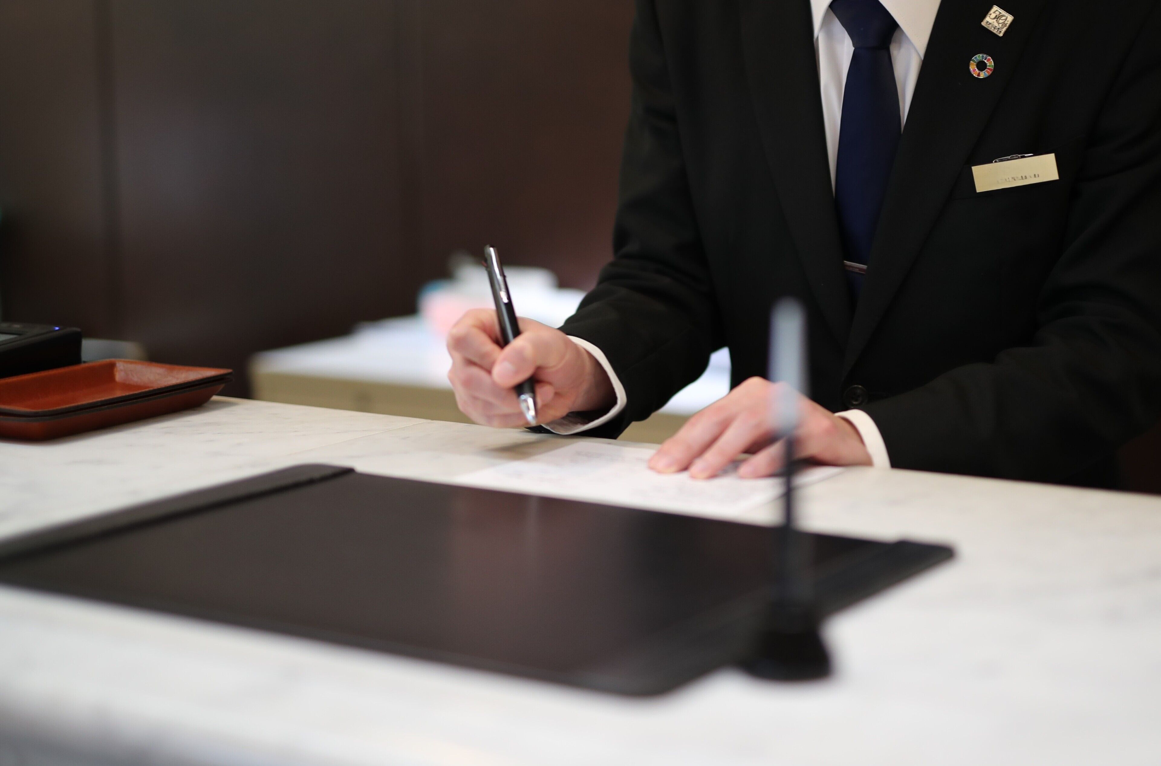 A person in a black suit with a name tag, holding a pen, and writing on a notepad at a desk with a tablet and a small orange tray.