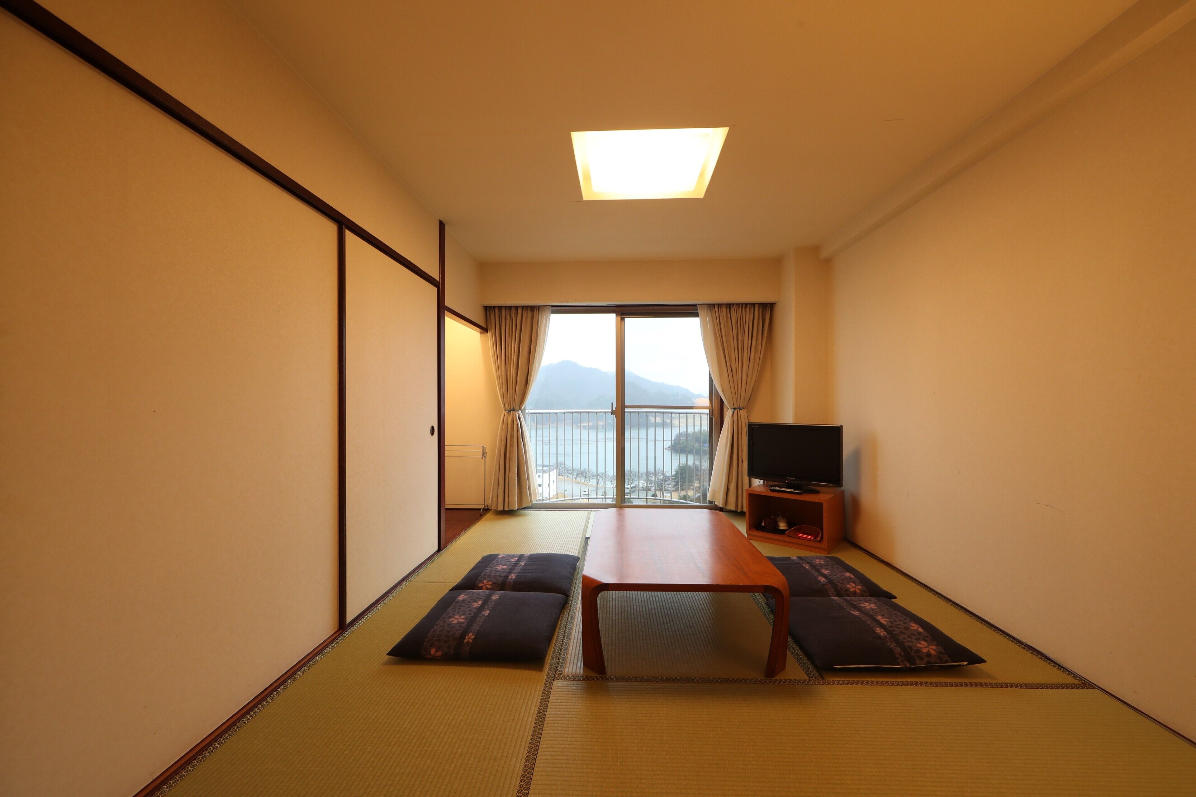 A hotel room with tatami flooring, a wooden table, two tatami mats, a television on a wooden stand, and a view of mountains through a sliding door.