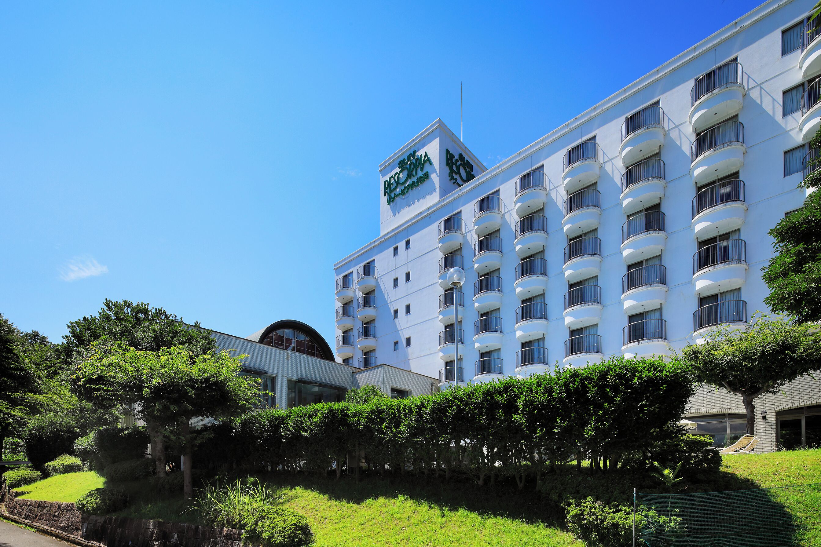 A multi-story hotel with balconies, a clear sky, and surrounding greenery.