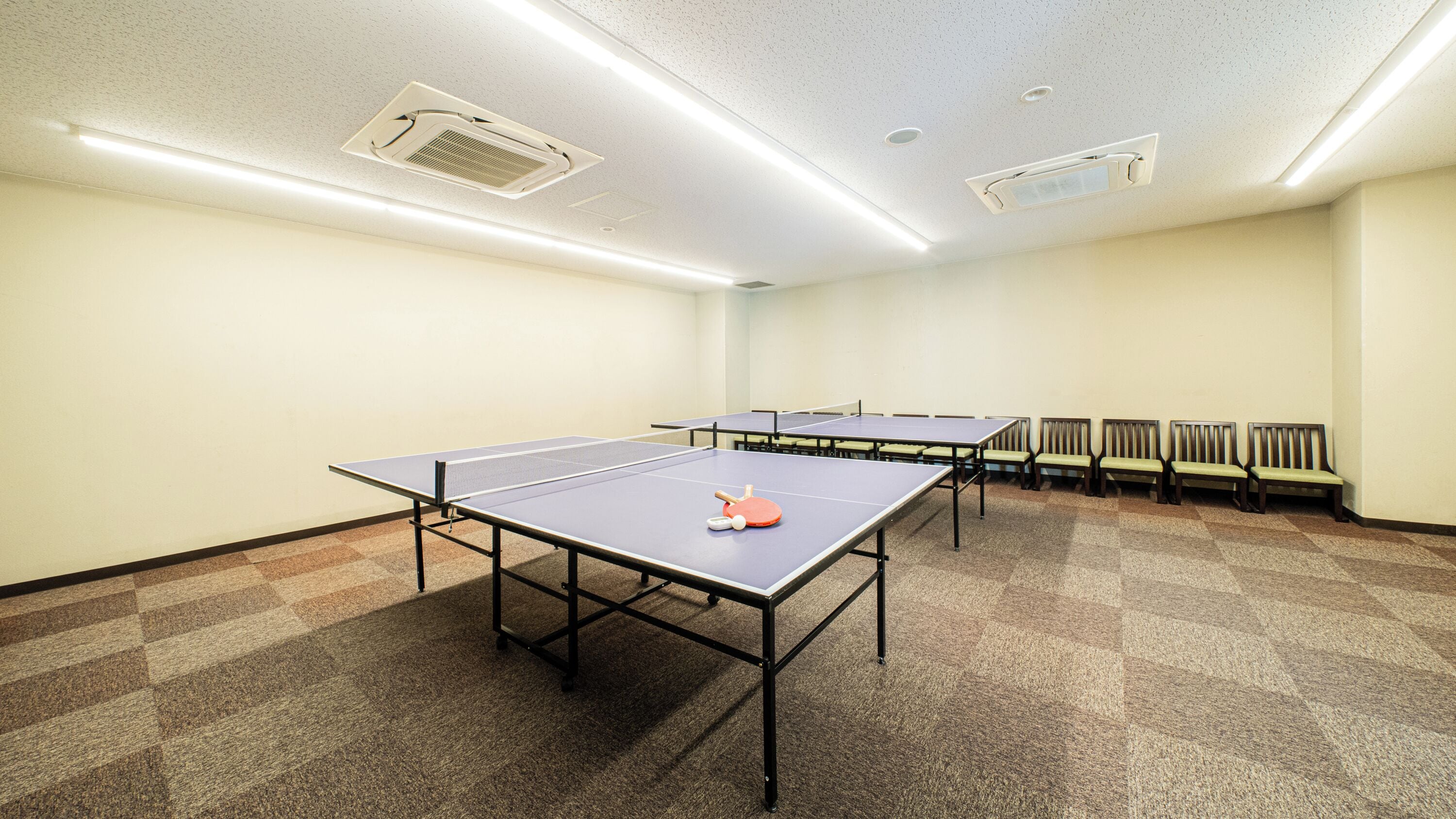 A room with ping pong tables, chairs, and ceiling lights.
