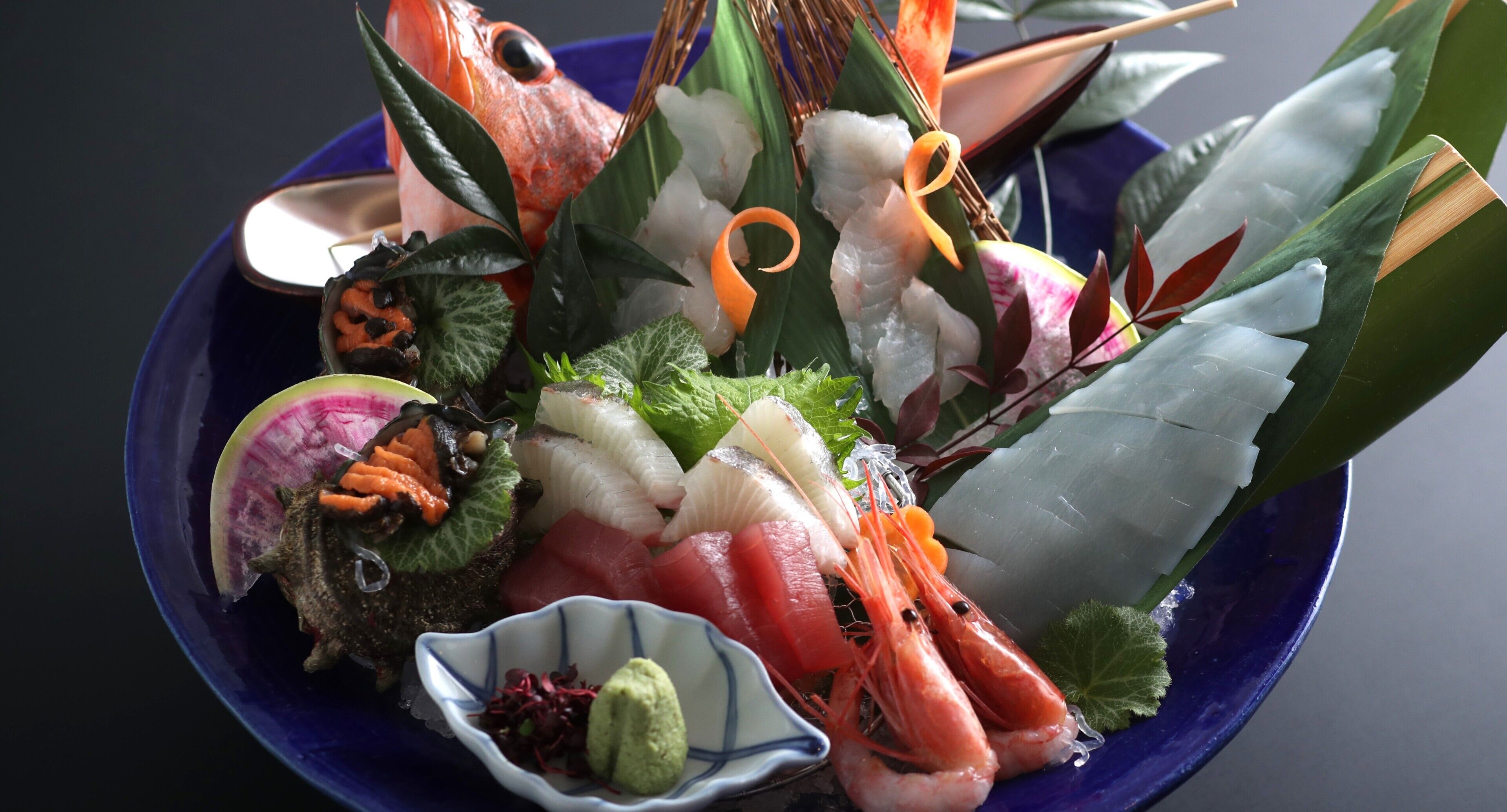 A bowl of assorted seafood including shrimp, fish, and other delicacies, with a small dish of green sauce.