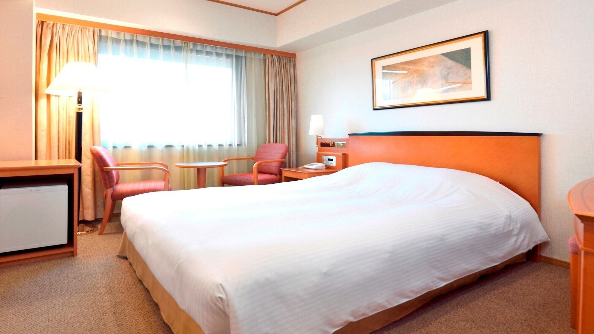 A hotel room with a large bed, a desk, two chairs, a lamp, a painting on the wall, and a window with curtains.