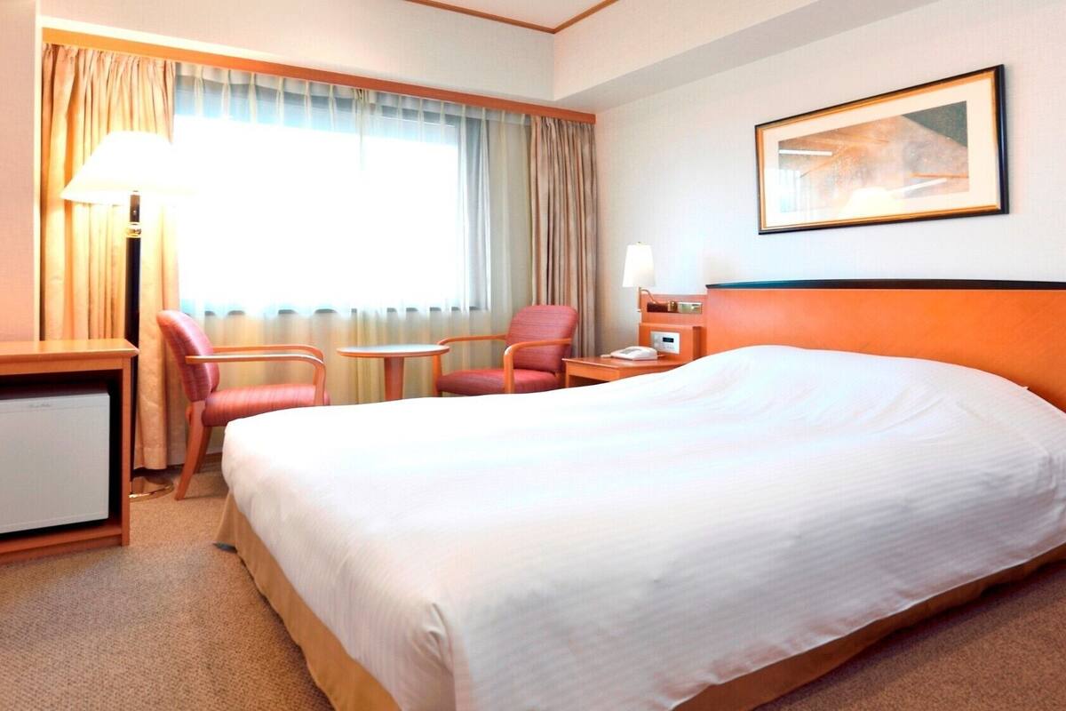 A hotel room with a large bed, a desk, two chairs, a lamp, a painting on the wall, and a window with curtains.