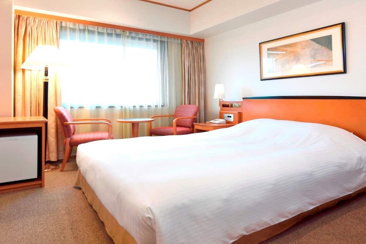 A hotel room with a large bed, a desk, two chairs, a lamp, a painting on the wall, and a window with curtains.