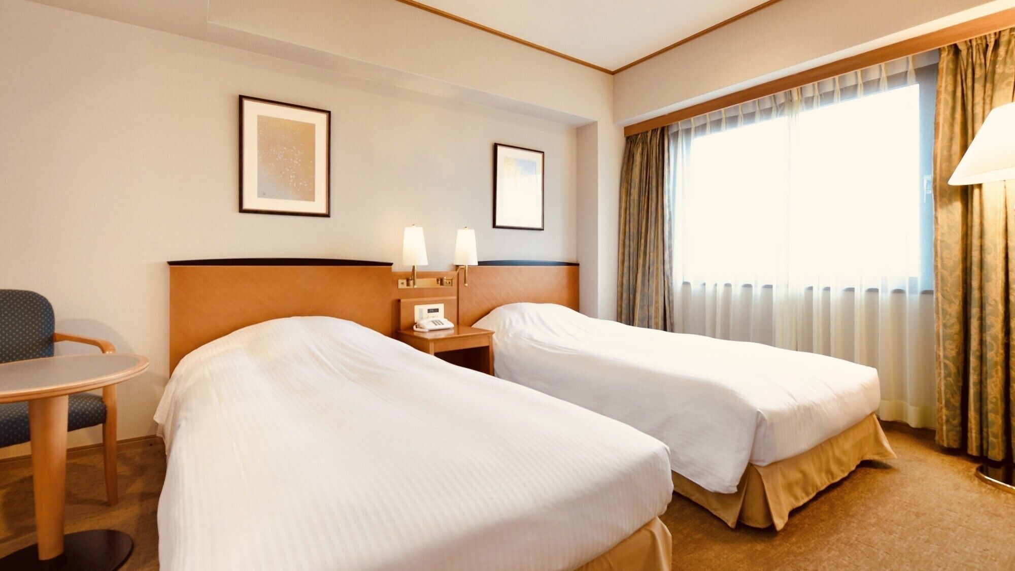 A hotel room with two beds, a small table, a chair, a nightstand with a lamp, and a window with curtains.