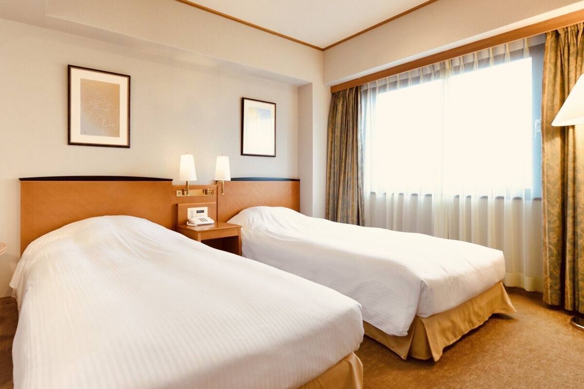 A hotel room with two beds, a small table, a chair, a nightstand with a lamp, and a window with curtains.