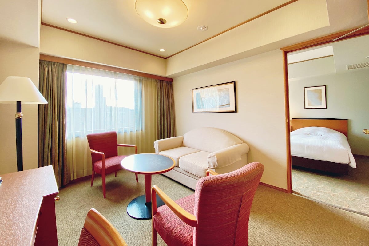A hotel room with a bed, a sofa, a small table, a chair, a lamp, and a window with curtains.