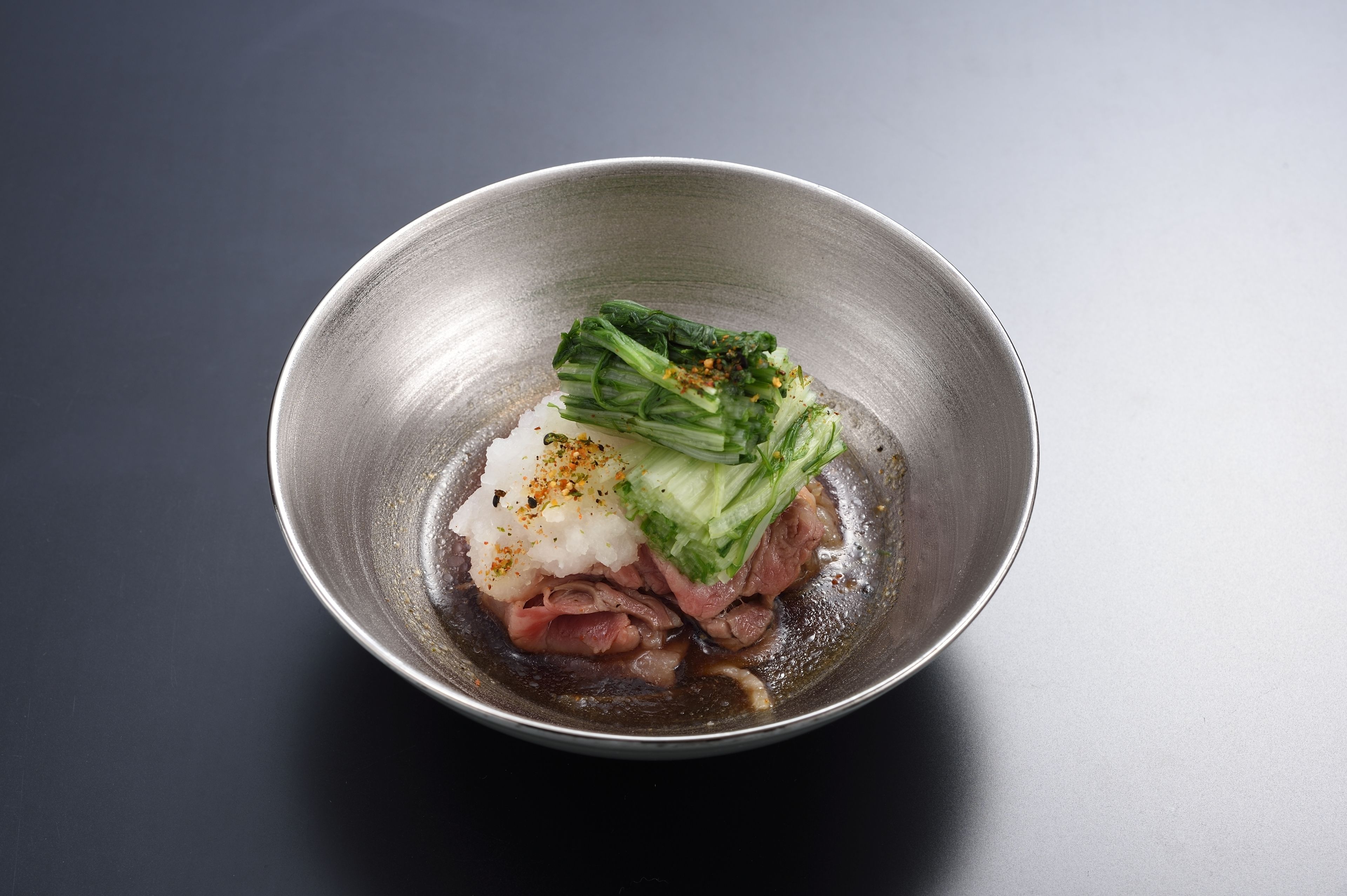 A metal bowl containing sliced meat, white rice, green vegetables, and a dark sauce.