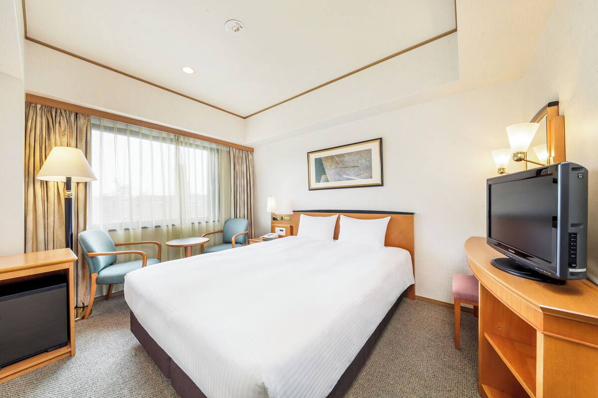 A hotel room with a large bed, a desk with a television, a chair, a small table, and a window with curtains.
