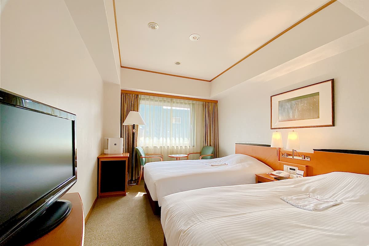 A hotel room with two beds, a TV, a desk with a lamp, and a window with curtains.