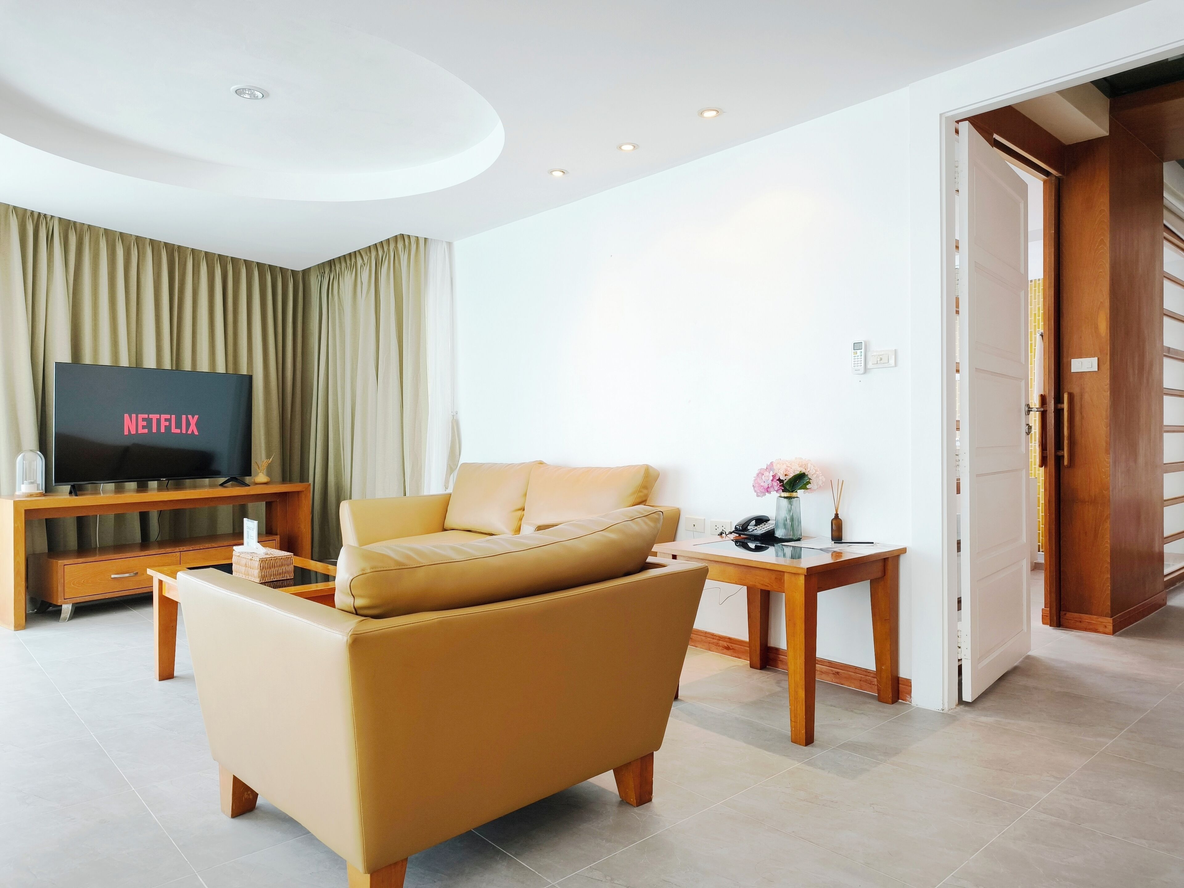 A modern living room with a sofa, a TV displaying 'NETFLIX', a small table with a vase, and a wooden door leading to another room.