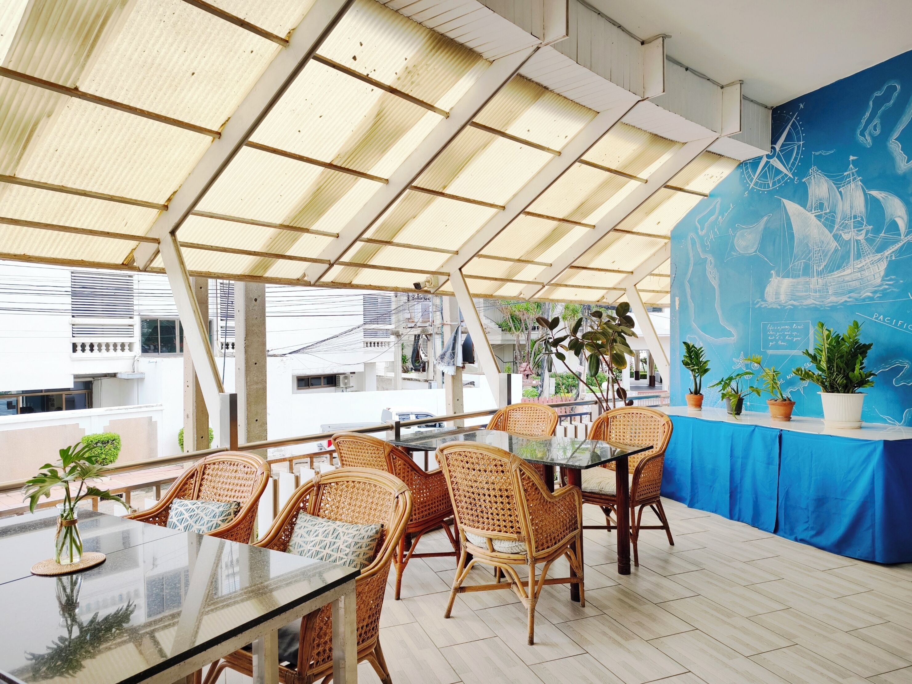 Uma área de refeições ao ar livre com cadeiras de vime e uma mesa de vidro, apresentando um mural azul com elementos náuticos e vista para edifícios ao fundo.