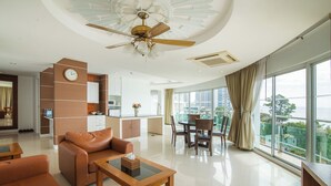 A modern living room with a sofa, coffee table, and dining area. A kitchen is visible in the background with a clock on the wall. - Royal Beach View Suites (Pattaya)