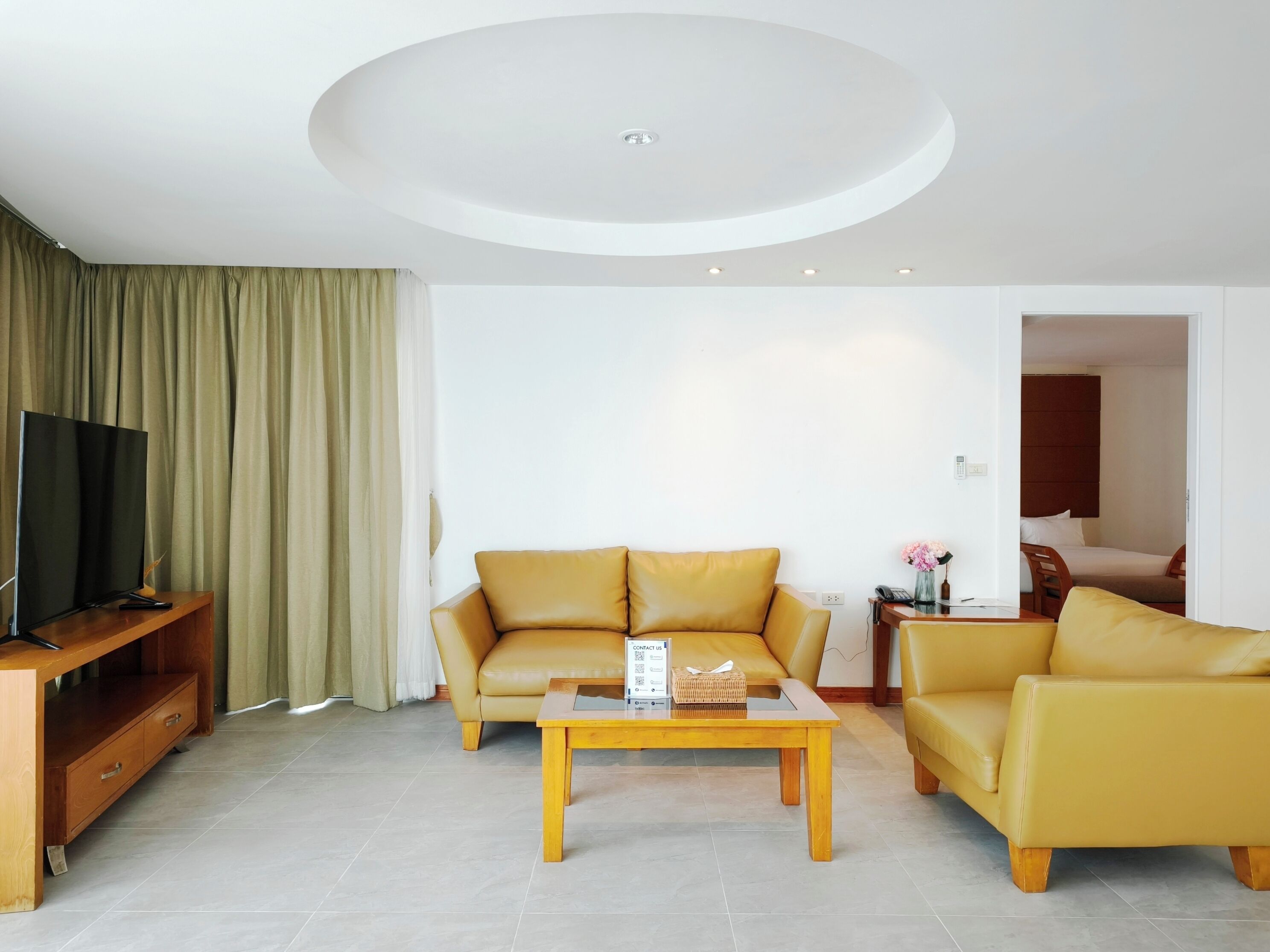 A modern living room with a sofa, armchair, and a coffee table. A TV is on a wooden stand, and a door leads to a bedroom.
