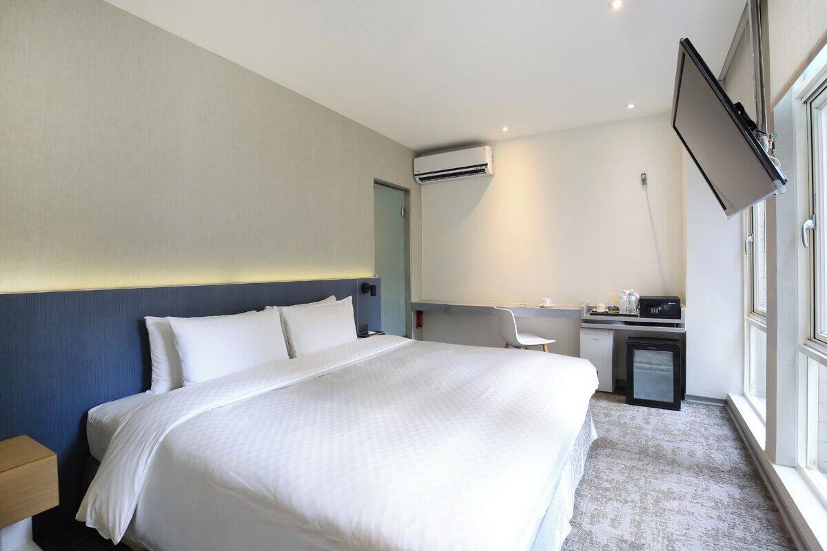 Ein Hotelzimmer mit Bett, Schreibtisch, Stuhl, Klimaanlage und einem an der Wand befestigten Fernseher.