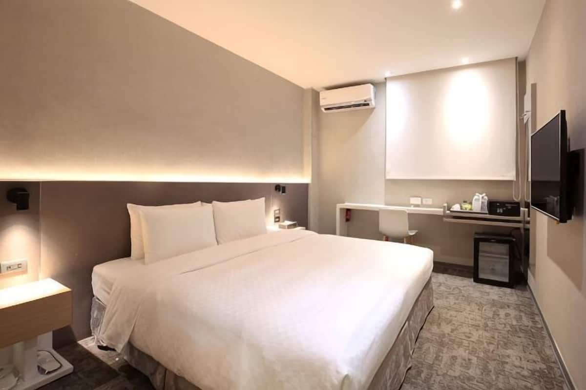 Ein Hotelzimmer mit einem großen Bett, einem Nachttisch, einem Fernseher, einer Klimaanlage und einem kleinen Schreibtisch mit Stuhl.
