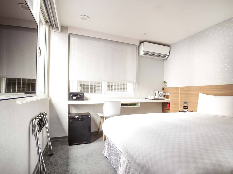 A hotel room with a bed, a desk, a chair, a radiator, and an air conditioning unit.